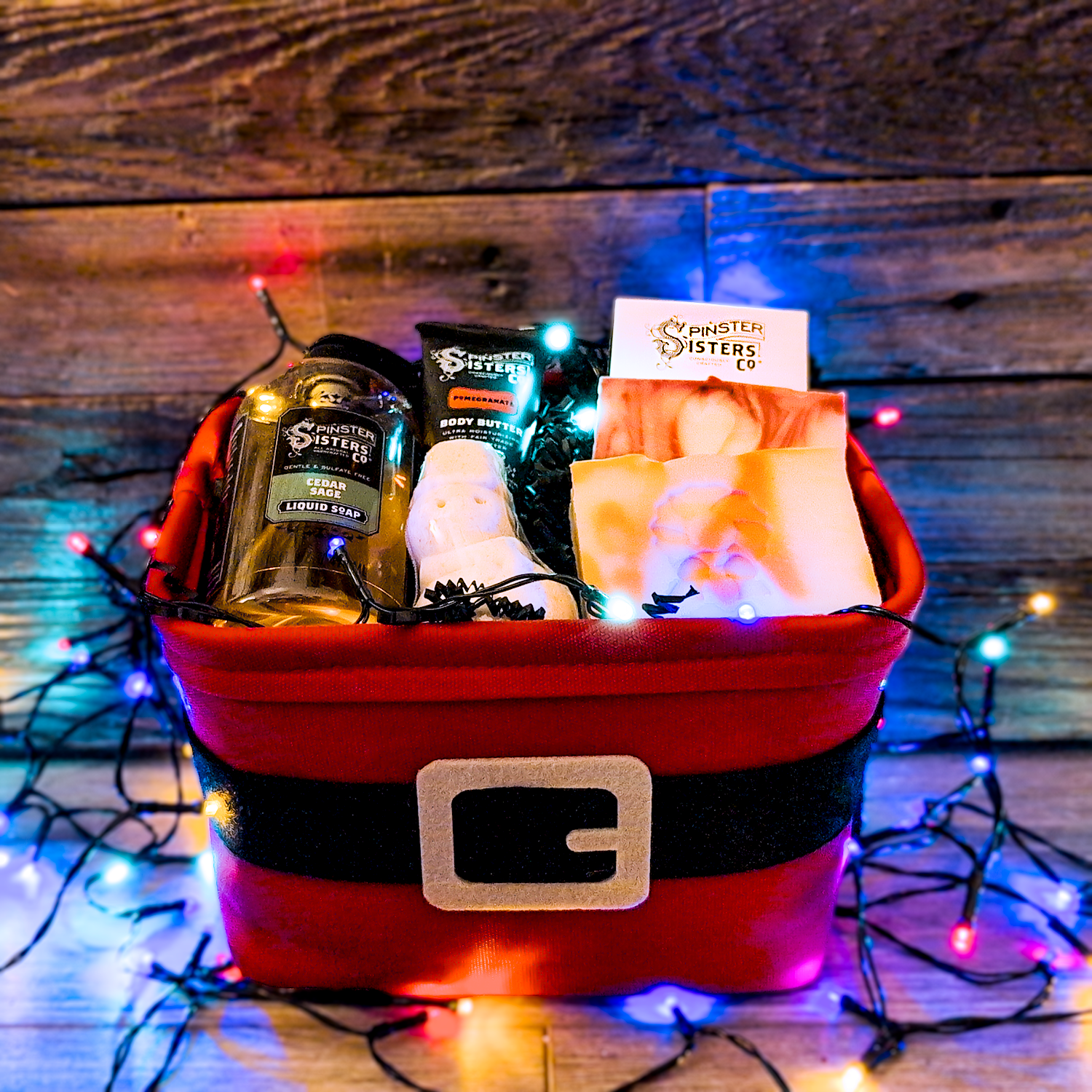 Santa Themed gift basket filled with Spinster Sisters Products on a wooden background with string lights.