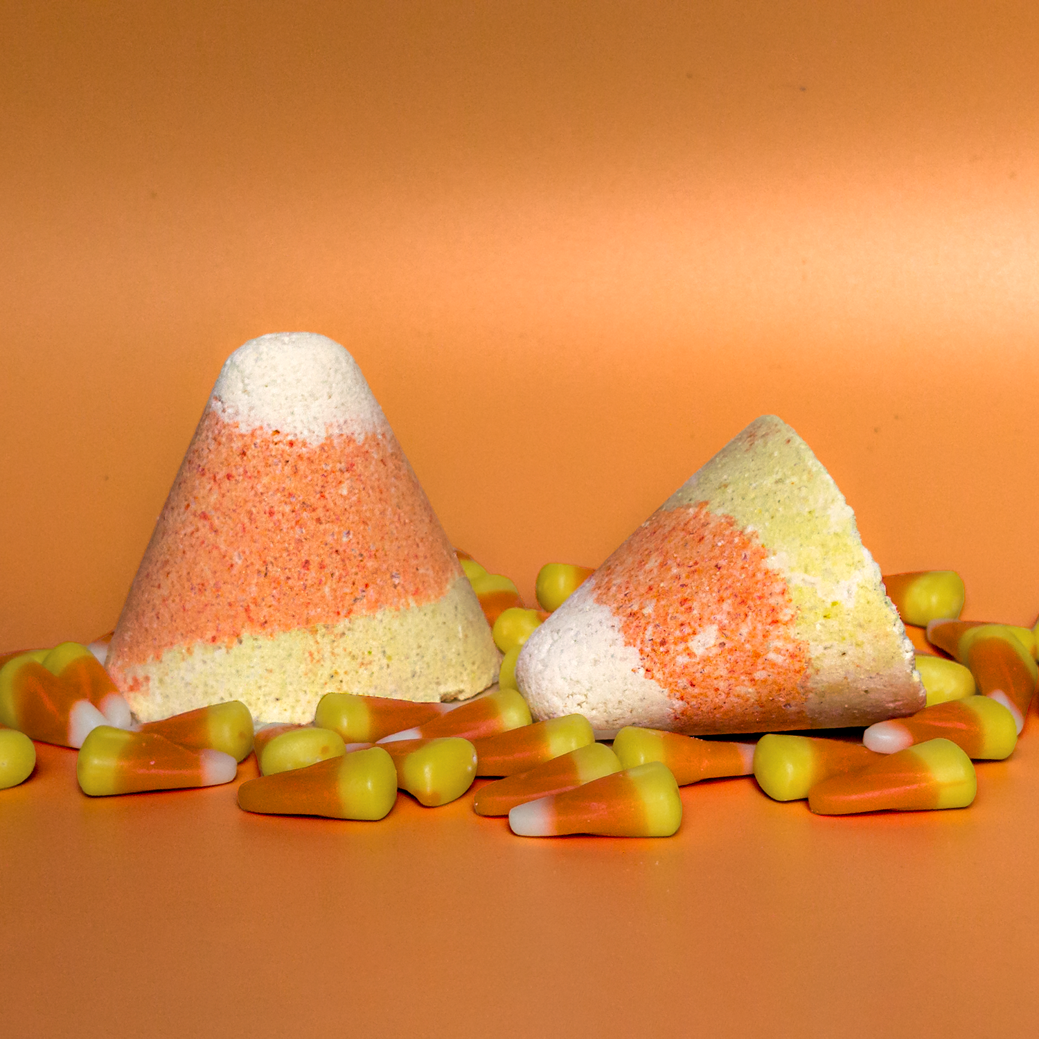 Two candy corn-themed bath bombs on an orange background with candy corn around them.