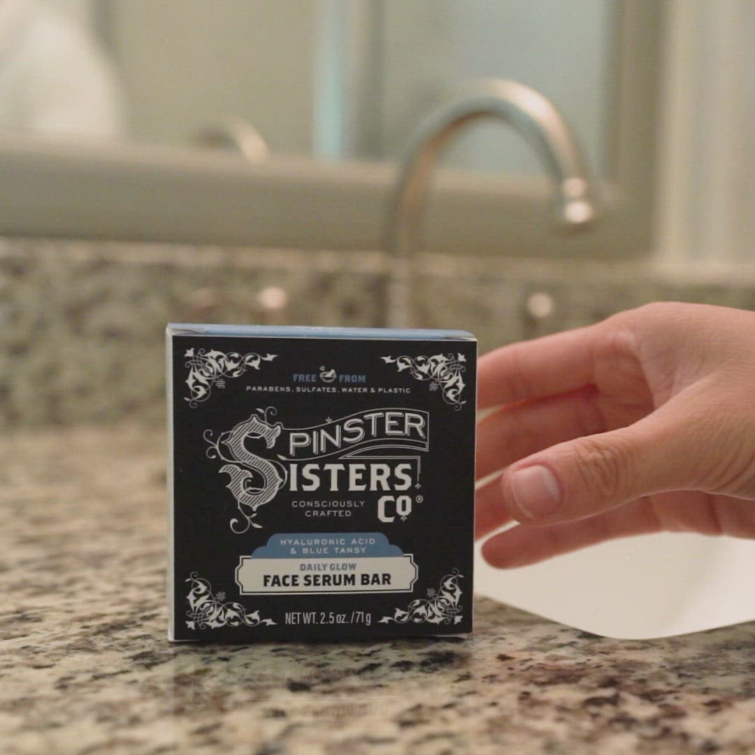 Spinster Sisters daily glow face face serum bar box on a counter. Someone picks up the blue bar and warms it in their hands and applies to face in a bathroom.Closes on Spinster Sisters logo (consciously crafted).