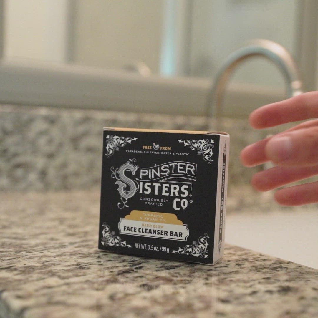 Spinster Sisters daily glow face cleanser bar box on a counter. Someone picks up the bright orange bar and washes their face in a bathroom sink. Follows by drying their face with a towel. Closes on Spinster Sisters logo (consciously crafted).